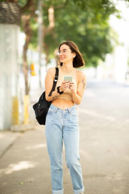 Cep telefonu ve el çantasıyla şehrin dışında yürüyen ve gözlerini kaçıran şık genç bir kadının portresi.