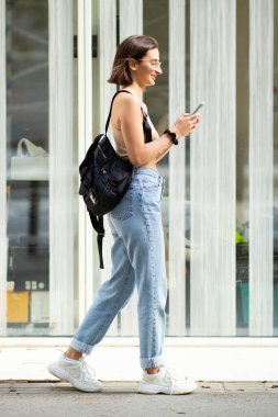 Şehirde çanta ve cep telefonuyla dolaşan genç bir kadının tam vücut profili.
