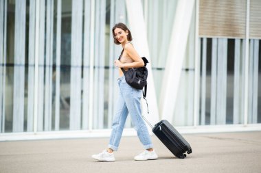 Gülümseyen genç bir kadının el çantası ve bavuluyla caddede yürüyüşünün tam boy portresi.