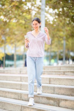 Kulaklıklı, merdivenlerden inip müzik dinlerken telefona bakan genç bir kadının vücut portresi.