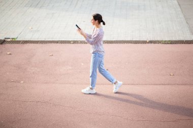 Kulaklık takan, cep telefonuyla mesajlaşan ve şehir dışında yürüyen genç bir kadının vücut portresi.