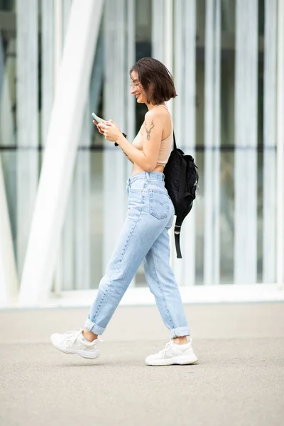 Full body profile portrait of stylish young woman walking with bag and mobile phone in the city