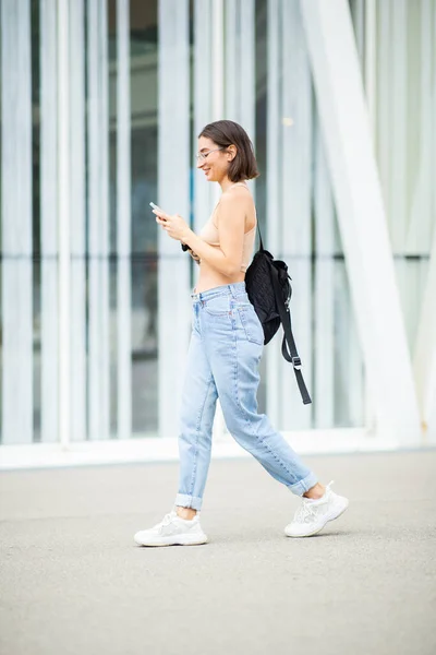 Full body profile portrait of beautiful young woman walking with bag and reading text message on her mobile phone in the city