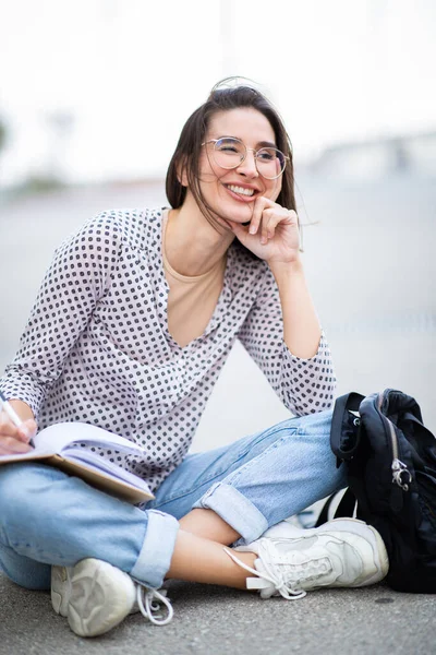 幸せな若い女性の肖像画で外に座ってペンと本を離れて見て 考えて — ストック写真
