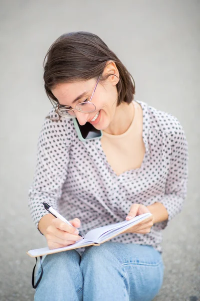 Портрет Счастливой Молодой Женщины Сидящей Снаружи Пишущей Книге Разговаривающей Мобильному — стоковое фото