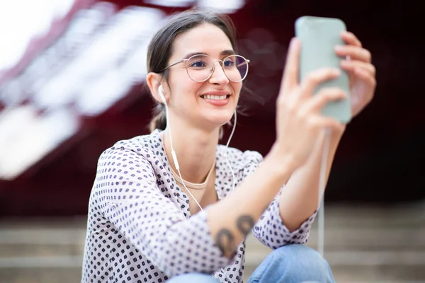 Close up side portrait of happy young woman holding cellphone and listening to music with earphones while sitting outside in the city