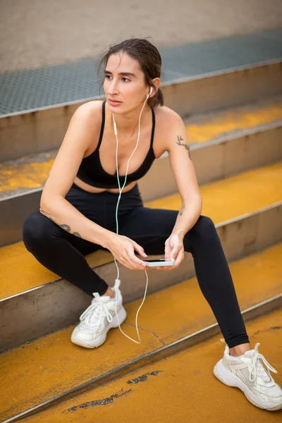 Портрет Фитнеса Молодой Женщины Сидящей Ступеньках Снаружи Наушниками Мобильным Телефоном — стоковое фото