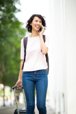Şehirde cep telefonuyla konuşurken bavulu çeken mutlu genç kadın.