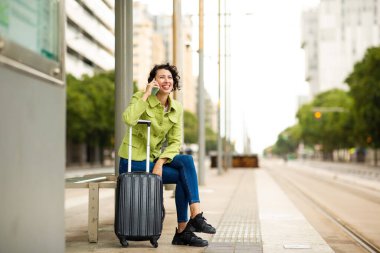Gülümseyen genç bir kadın valizin sapına yaslanıyor ve otobüs durağında beklerken cep telefonuyla konuşuyor.