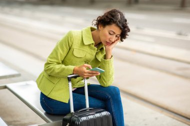 Şehirdeki otobüs durağında bavuluyla beklerken cep telefonu kullanan üzgün genç bir kadın.
