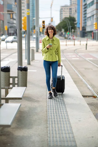 Happy woman pulling rolling suitcase and text messaging using mobile phone outdoors in city
