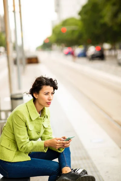 Piękna Młoda Kobieta Czeka Przystanku Autobusowym Pomocą Telefonu Komórkowego Zewnątrz — Zdjęcie stockowe