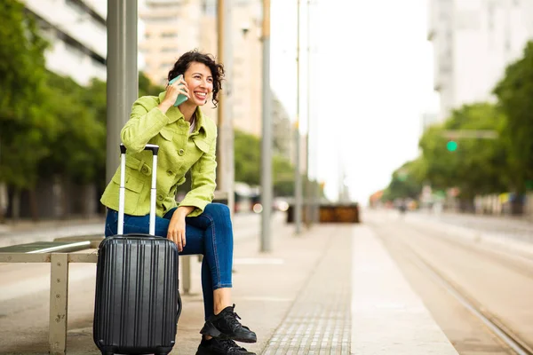 在城市里 年轻快乐的女人靠在手提箱上 一边用手机聊天 一边在巴士站等着 — 图库照片
