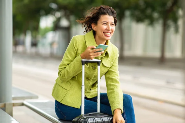Cheerful young woman with luggage and mobile phone outdoors at city bus stop