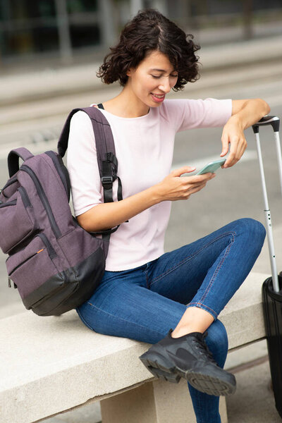 Happy Female Traveler Messaging Using Mobile Phone While Sitting Outdoors Stock Photo