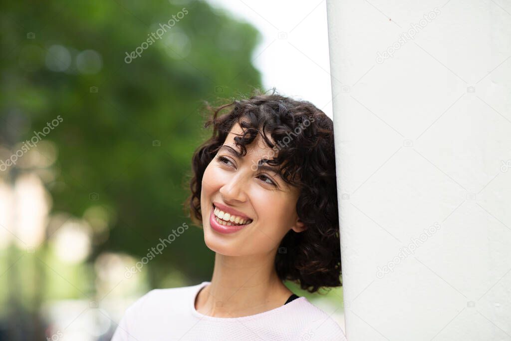 Cheerful young woman leaning on wall and looking away in thoughts