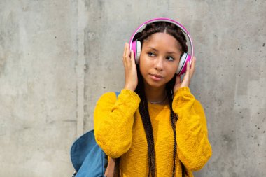 Gri duvarda duran, müzik dinlemek için kulaklık takan Afrikalı genç bir öğrencinin portresi.