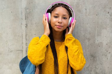 Gri duvarın yanında durup kulaklıkla müzik dinleyen Afrikalı genç bir kız öğrencinin portresi.