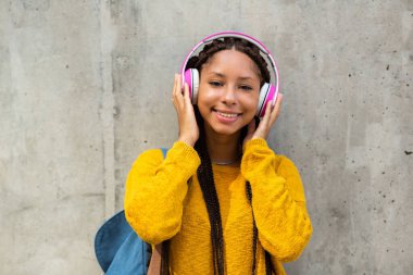 Gri duvarın yanında durup kulaklıkla müzik dinleyen gülümseyen zenci kız öğrencinin portresi.