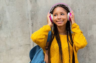 Yüzünde çanta ve kulaklıklarla duvarın önünde dikilen gülümseyen Afrikalı genç öğrencinin portresi.