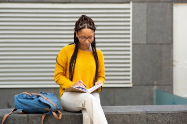Genç Afro-Amerikan kız öğrencinin portresi. Dışarıda oturmuş kitap yazıyor.