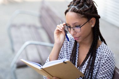 Genç Afro-Amerikan kadının yakın plan portresi. Gözlüğünü ayarlarken dışarıda oturup kitabına bakıyor.