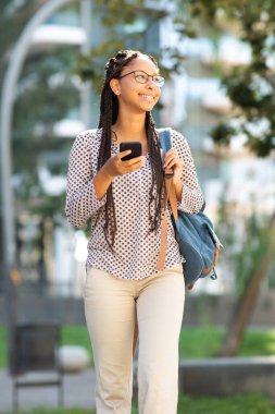 Dışarıda cep telefonuyla yürüyen gözlüklü mutlu Afro-Amerikalı bir kızın portresi.