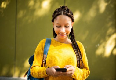 Sarı duvarın yanında dururken cep telefonuna bakan gülümseyen Afrikalı bir kadının portresi.