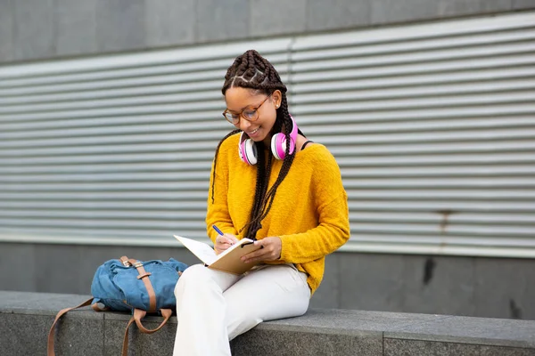 Gülümseyen Afrikalı genç bir kadının portresi. Elinde çantasıyla dışarıda oturuyor. Not defterine yazıyor.