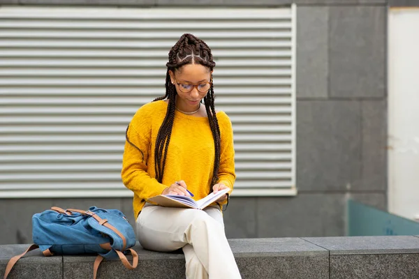 Genç Afro-Amerikan kız öğrencinin portresi. Dışarıda oturmuş kitap yazıyor.