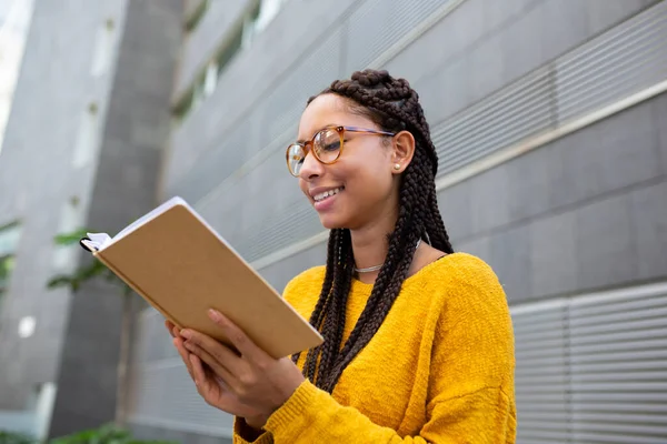Saçları örgülü, gözlüklü, kampüste kitap okuyan gülümseyen Afrikalı Amerikalı bir kızın portresi.