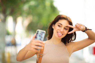 Barış işareti ile gülümseyen genç bir kadının portresi ve dışarıda cep telefonuyla selfie çekmesi.