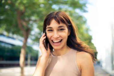 Şehirde kameraya bakıp cep telefonuyla konuşan neşeli genç bir kadının portresini kapat.