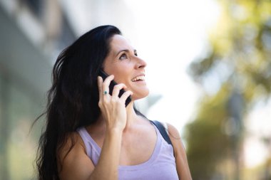 Neşeli beyaz kadın cep telefonuyla konuşurken yukarı bakıyor.