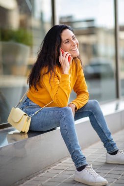 Şehirde dışarıda otururken rahat kot pantolon ve cep telefonuyla konuşan mutlu bir kadın.