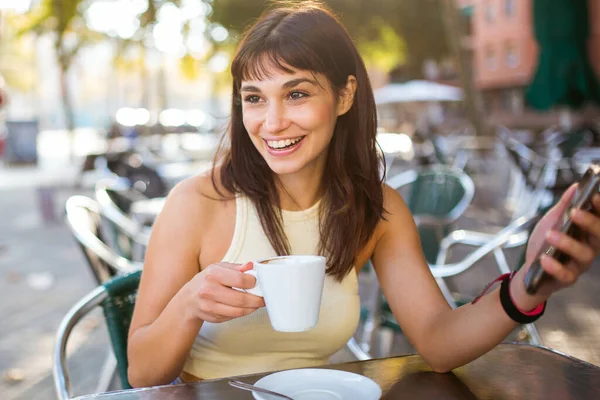 Portrait of beautiful young woman sitting at outdoor cafe with phone drinking cup of coffee