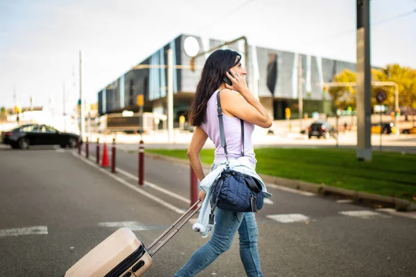 Profile of happy caucasian woman pulling rolling suitcase and carrying handbag while talking on mobile phone outdoors in city