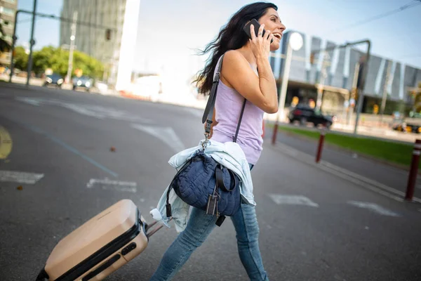 城市里 一个快乐的女人在户外用手机提着滚动的手提箱 提着手提包的照片 — 图库照片