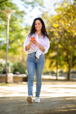 Parkta yürürken gülümseyen olgun bir kadın cep telefonu kullanıyor.