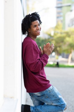 Gülümseyen Afro-Amerikan bir adam. Duvara yaslanmış poz veriyor.