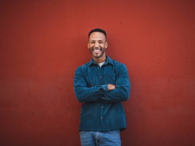 Neşeli, Afro-Amerikan bir adam. Kollarını kestane rengi duvara dayamış poz veriyor.