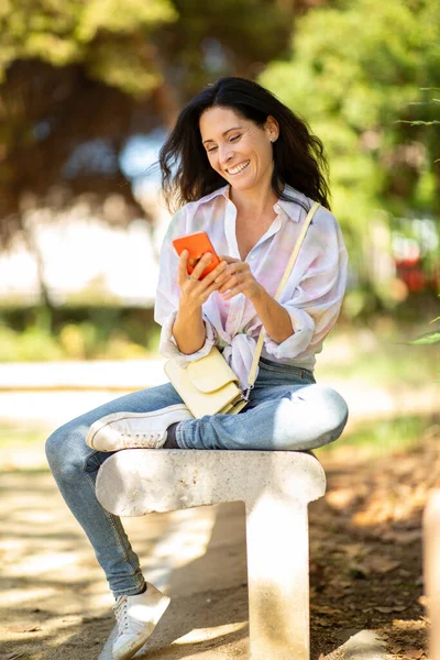 公園のベンチで屋外の携帯電話を使用して笑顔の白人女性のテキストメッセージ — ストック写真