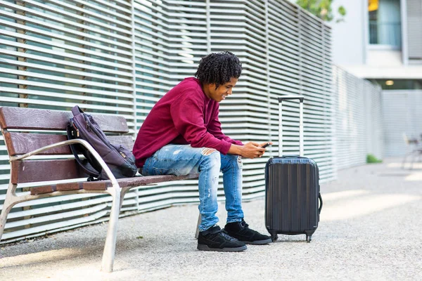 Czarny Mężczyzna Podróżujący Pomocą Smartfona Podczas Siedzenia Ławce Mieście Bagażem — Zdjęcie stockowe