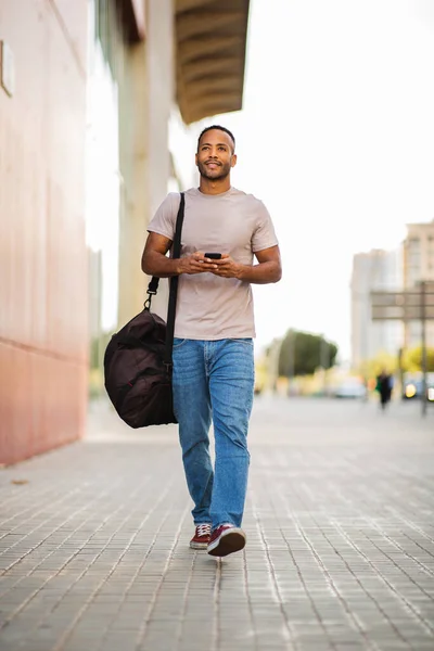 Young african american man with smart phone and bag walking outdoors in city