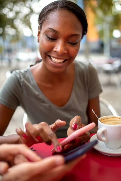 Happy african american woman using mobile phone while sitting with her friend outdoor at coffee shop