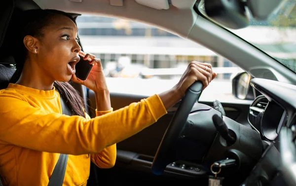 Young black woman shocked while talking on mobile phone and driving car outdoors