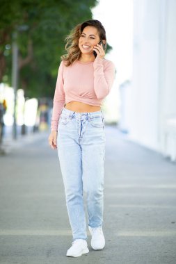 Gülümseyen genç kadının cep telefonuyla yürüyüp konuşmasının tam uzunlukta portresi