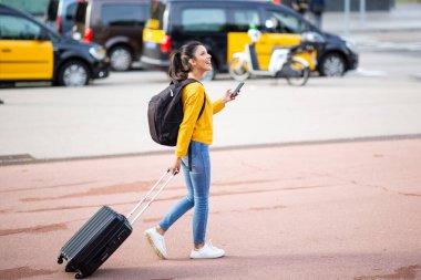 Şehirde cep telefonuyla valiz taşıyan mutlu genç bir kadının tam boy portresi.