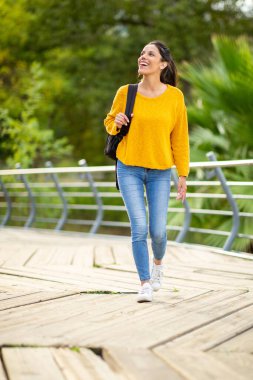 Dışarda çantasıyla yürüyen gülümseyen genç bir kadının tam boy portresi