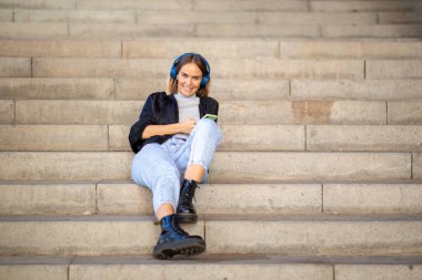 Merdivenlerde dinlenen, cep telefonu ve kulaklıkla müzik dinleyen gülümseyen kadın portresi. 